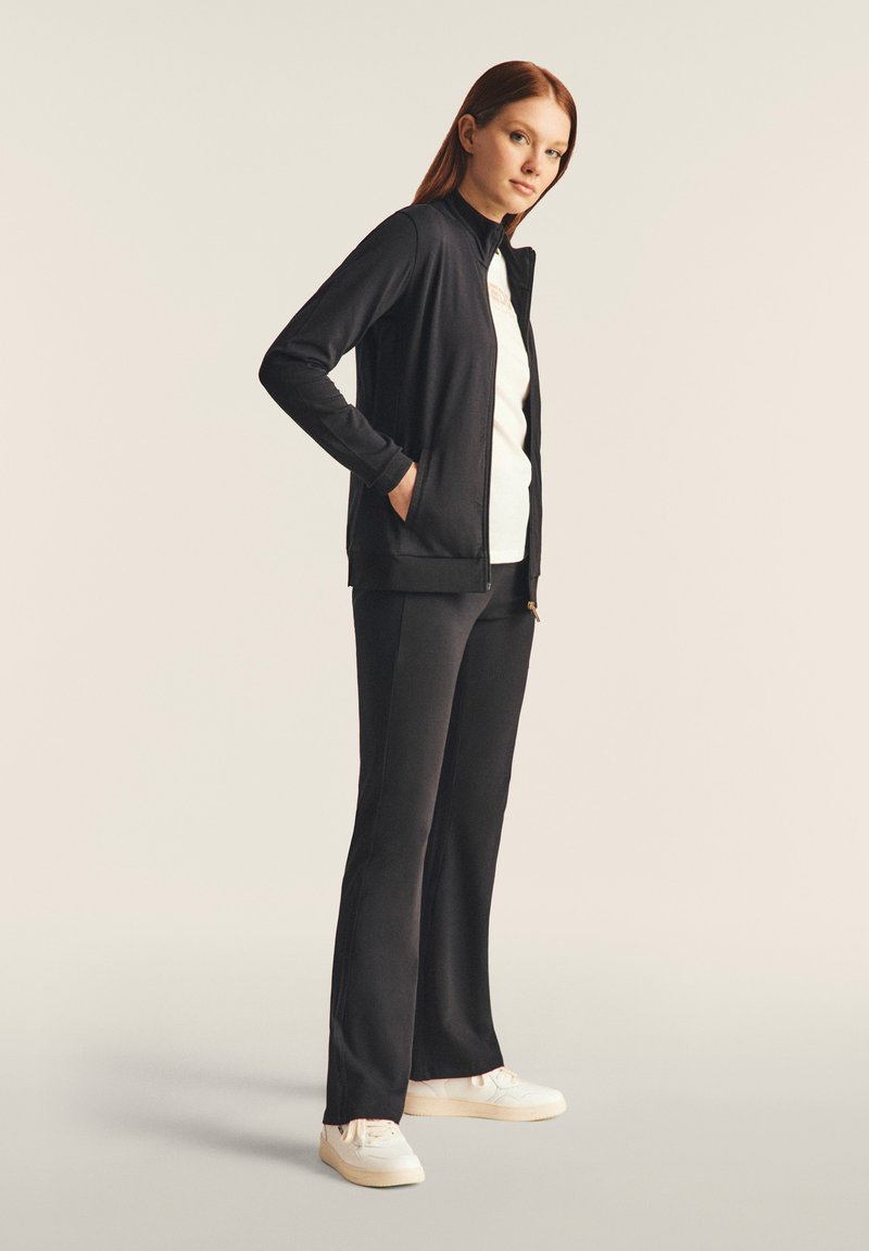 Chaqueta deportiva negra con cuello alto, cierre de cremallera y puños acanalados, combinada con pantalones negros de campana y zapatillas blancas. Textura suave.