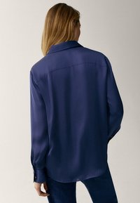 Donna con capelli biondi che indossa una blusa blu scuro a maniche lunghe e larghe, e pantaloni scuri, voltata di spalle contro uno sfondo neutro.