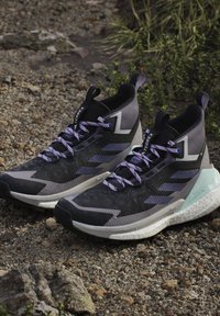 Paire de chaussures de trail montantes noires et violettes avec des accents bleu clair sur un sol rocailleux en extérieur près de plantes vertes.