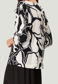 Blouse en coton avec un motif floral noir et blanc, des manches trois-quarts et une coupe décontractée, assortie à une jupe plissée noire.