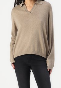Pull léger beige avec un décolleté en V et un col. Coupe ample, manches longues et ourlet côtelé. Associé à un pantalon noir ajusté.