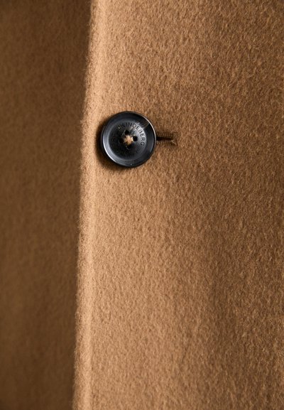 Bruine wollen stof met een zachte textuur, met een zwarte knop met een kruisontwerp aan de voorkant bevestigd.