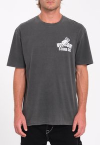 Camiseta de algodón gris con mangas cortas, escote redondo y un logo blanco impreso en el lado izquierdo. Textura suave, corte casual.