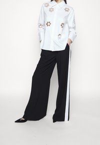 Vit skjorta med blommig broderi, tillsammans med svarta vida byxor med en vit sidostrip. Skorna är svarta spetsiga ballerinas.