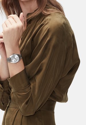 Femme portant un chemisier en satin vert olive montrant une montre-bracelet argentée avec un bracelet en métal à son poignet gauche sur un fond clair.