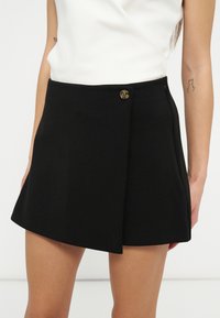 Femme portant une jupe portefeuille noire avec une fermeture à bouton unique à la taille et un haut blanc sans manches.