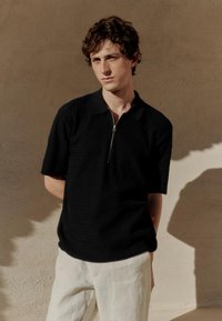 Mladi moški z kodrastimi lasmi, oblečen v črno teksturirano kratko majico z zadrgo in svetle hlače, stoji ob bež steni.