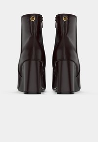 Pair of dark brown leather ankle boots with block heels and gold circular logos on the back zipper pulls, shown from the rear.