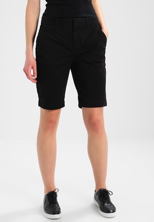 Person iført sorte knælange shorts, sort top og sorte sneakers, der står mod en ensartet lys baggrund.