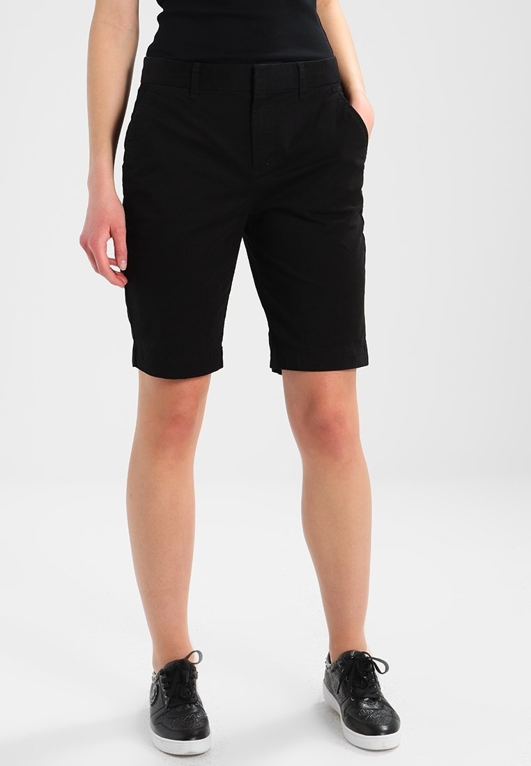 Personne portant un short noir mi-cuisse, un haut noir et des baskets noires, debout devant un fond clair uni.