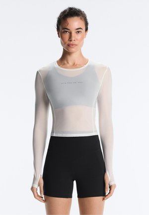 Femeie care poartă un top crop alb transparent cu mâneci lungi cu textul „TU CONTRA TU” și pantaloni scurți negri cu talie înaltă, stând pe un fundal simplu.