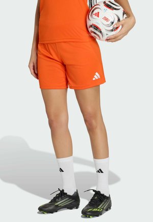 Osoba ubrana w pomarańczowe spodenki i koszulkę piłkarską, białe skarpety Adidas oraz czarne korki, trzymająca biało-czerwoną piłkę piłkarską Adidas.