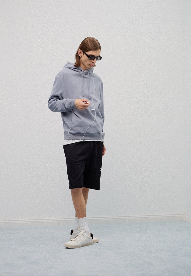 Jasnoszara bluza z kapturem i sznurkiem, czarne szorty, białe skarpetki i kremowe sneakersy. Noszący okulary przeciwsłoneczne i trzymający urządzenie z słuchawkami.