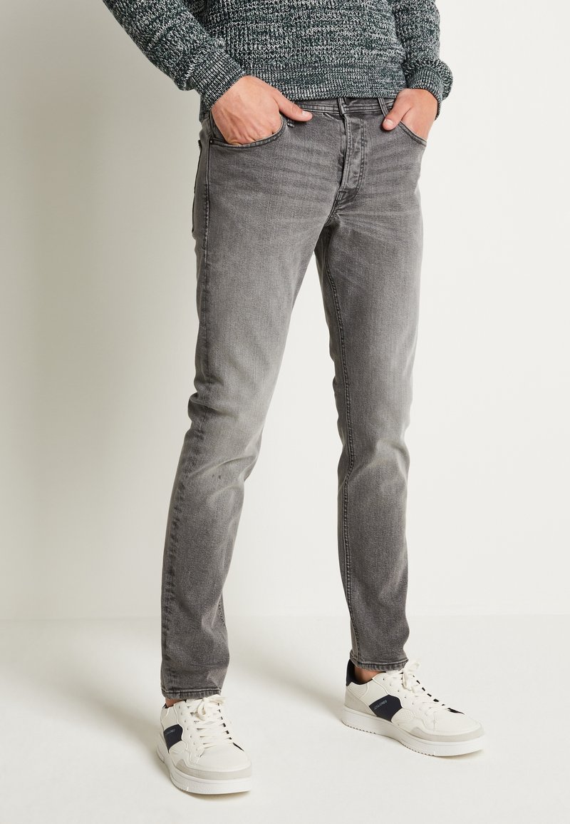 Homme portant un jean gris délavé, des baskets blanches et un pull en maille gris, les mains dans les poches devant un fond uni.