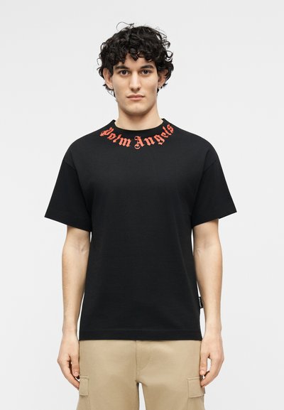 Göndör hajú férfi fekete pólóban, amelynek nyakánál piros "Palm Angels" felirat, és bézs színű cargo nadrágot visel, egyszínű háttér előtt.