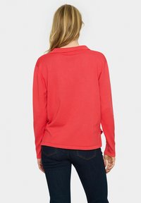 Dlhý rukáv, koralová polo košeľa s golierom, rebrovanou textúrou a bočnými rozparkami, v kombinácii s tmavými džínsami. Viditeľné švy na švoch.