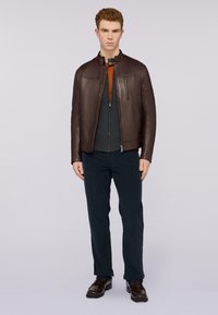 Chaqueta de cuero marrón con cuello alzado, suéter gris con cremallera debajo y pantalones oscuros. Modelo con zapatos oscuros se encuentra contra un fondo simple.