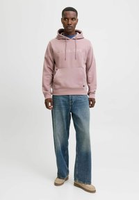 Helle rosa Kapuzenjacke mit Vordertasche, Kordelzug und Logo. Kombiniert mit lockeren blauen Jeans und beigen Schuhen. Glatte Textur.