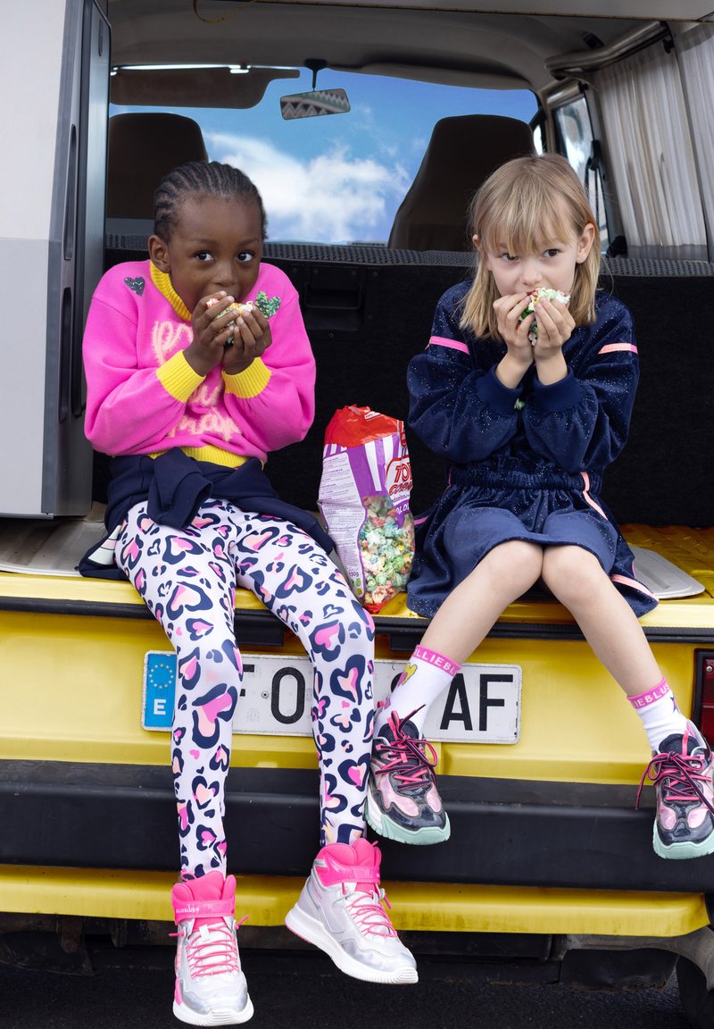 Twee kinderen zitten op de achterkant van een gele bestelwagen. De een draagt een roze trui en gestreepte leggings; de ander draagt een donkere outfit. Beiden eten snacks.