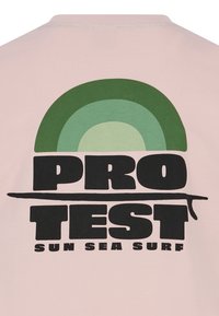Světle růžové tričko s barevným kruhovým grafikou v zelené a modré, se silným černým textem: "PRO TEST SUN SEA SURF."