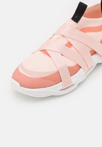 Baskets rose clair avec des sangles élastiques croisées, dessus en tricot, languette noire et semelle blanche épaisse sur fond blanc.