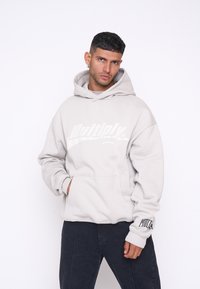 Felpa con cappuccio grigio chiaro, dotata di tasca frontale e grande logo bianco. Presenta polsini a costine e una vestibilità rilassata. Pantaloni neri per contrasto.