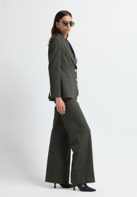 Donkergroene pak met een getailleerde blazer met gouden knopen en wijde broekspijpen. Het model draagt reflecterende zonnebrillen en zwarte hakschoenen.