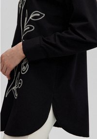 Black shirt with white floral embroidery, long sleeves, and a curved hem. Side slit details enhance movement; fabric appears soft and smooth.