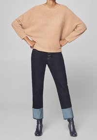 Beige oversized gebreide trui met een brede halslijn, gecombineerd met donkerblauwe jeans met lichtblauwe omslagzomen en enkellaarsjes.