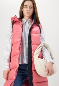 Gilet smanicato imbottito di un vivace rosa con cappuccio, bordi neri e finitura lucida. Modello che indossa una camicia a righe con bottone e una borsa color crema.