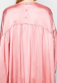 Back view of a person wearing a loose, shiny pink blouse with fabric-covered buttons on the upper back and gathered detailing below the yoke.