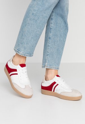 Osoba nosząca białe i czerwone trampki z beżowymi podeszwami i jasnoniebieskie dżinsy na jednolitym jasnym tle.