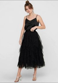 Black tulle skirt with layered ruffles, worn with a sleek black satin top and clear block-heeled sandals. Smooth texture, mid-calf length.