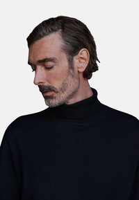 Maglione nero scuro a collo alto con texture a coste, caratterizzato da un colletto alto e una vestibilità rilassata, indossato da un uomo con capelli di lunghezza media.