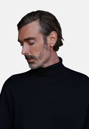 Maglione nero scuro a collo alto con texture a coste, caratterizzato da un colletto alto e una vestibilità rilassata, indossato da un uomo con capelli di lunghezza media.