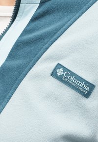 Κοντινό πλάνο γαλάζιου και τιρκουάζ φλις μπουφάν με φερμουάρ, με κεντημένη ετικέτα λογότυπου της Columbia Sportswear Company στο στήθος.