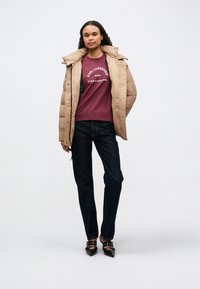 Abrigo acolchado en color beige claro, camiseta burdeos con texto blanco, jeans azul oscuro y zapatos de tacón negros con detalles metálicos.