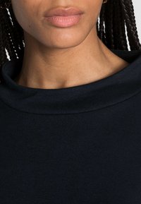 Mörk marinblå topp med en slät, mjuk textur och en bred, vikbar halsringning. Närbild av kragen och den övre bröstområdet.