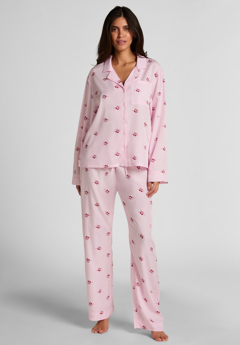 Σετ πιτζάμες σε απαλό ροζ ύφασμα, με τοπ κουμπωτό με τσέπη και μακριά μανίκια, συνδυασμένο με άνετο παντελόνι με μικρά κόκκινα λουλούδια.