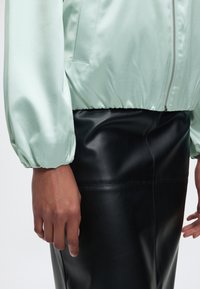 Lichtgroene satijnen jas met ritssluiting over een zwarte leren rok, getoond van taille tot halverwege de dij, met de handen van het model ontspannen langs het lichaam.