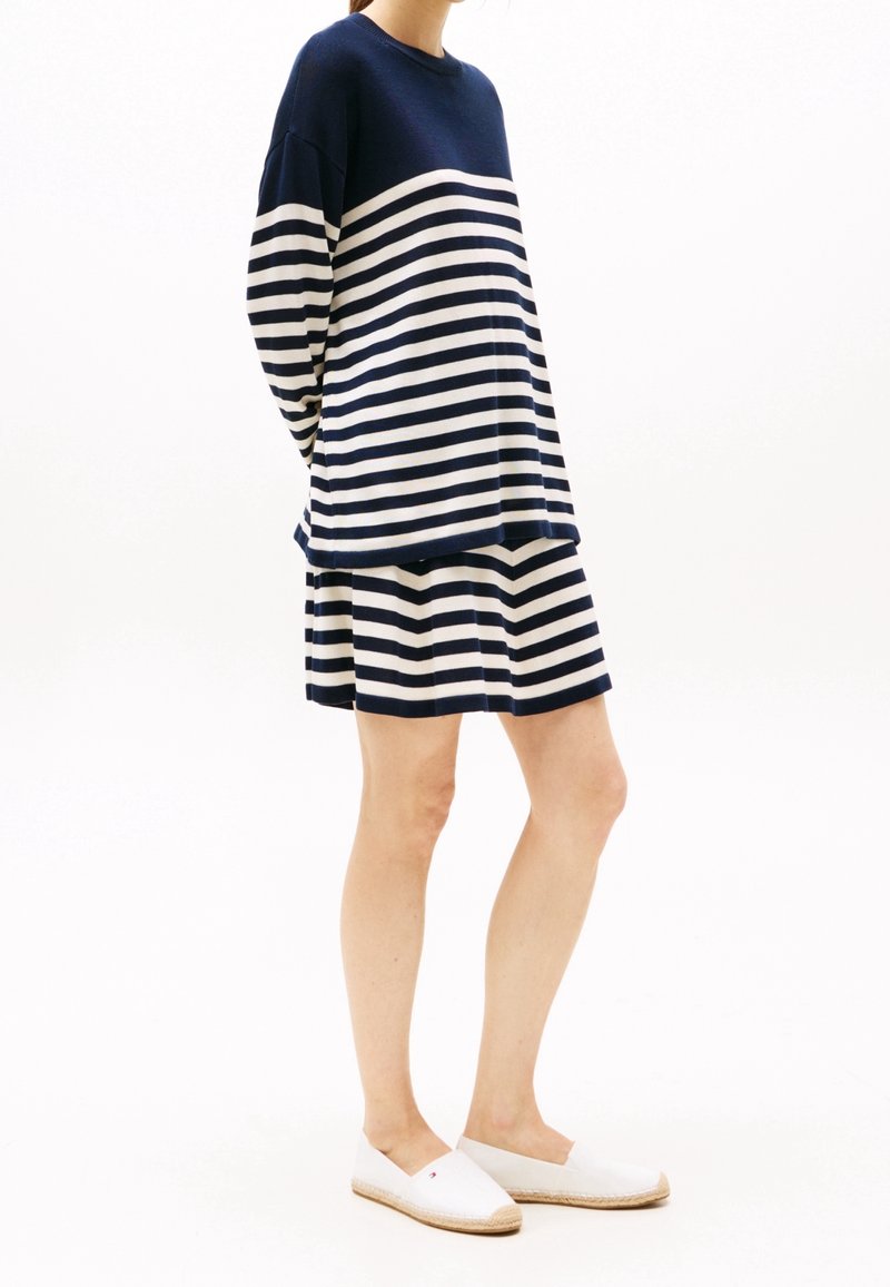 Femme portant un haut à manches longues rayé horizontalement bleu marine et blanc, avec une jupe assortie, associé à des espadrilles blanches slip-on.