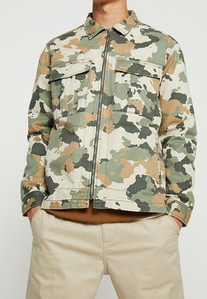 Mand iført en beige, grøn og brun camouflage-mønstret lynlåstrøje med to brystlommer med klap og beige bukser.