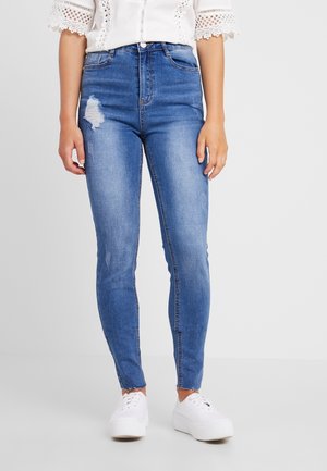 Kobieta ubrana w niebieskie dopasowane dżinsy z wysokim stanem z przetarciami, w połączeniu z białą koronkową bluzką z krótkim rękawem oraz białymi sneakersami na platformie.