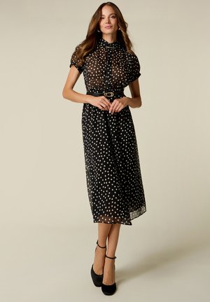 Woman wearing a black sheer midi dress with white polka dots, short sleeves, a belt, and black ankle-strap heels standing against a neutral background.