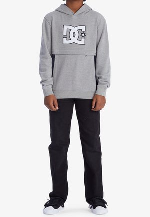 Sudadera gris con logo negro de DC, bolsillo tipo canguro y puños acanalados. Combinada con jeans negros y zapatillas negras. Estilo simple y casual.