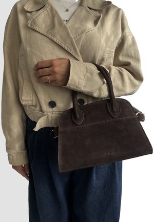 Borsa in suede marrone con manici arrotondati e chiusura con zip. Indossata con una giacca beige e pantaloni in denim scuri e larghi. Texture morbide.