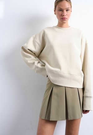 Jonge vrouw die een oversized crèmekleurige sweatshirt en een kaki geplooide mini-rok draagt, poserend met één hand op haar heup tegen een witte achtergrond.