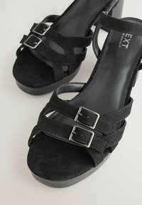 Crne suede sandale s visokom petom s dvostrukim kopčama, otvorenom nožnom prstom i teksturiranom platformom. Glatka unutarnja obloga i elegantan dizajn.