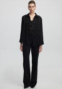 Camicia nera trasparente con bottoni indossata sopra un top in pizzo nero, abbinata a pantaloni neri a vita alta e tacchi a punta.