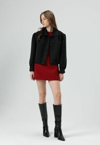 Zwarte cropped jas met pofmouwen, rode getailleerde jurk eronder en zwarte laarzen met hak tot aan de knie, tegen een lichtgrijze achtergrond.